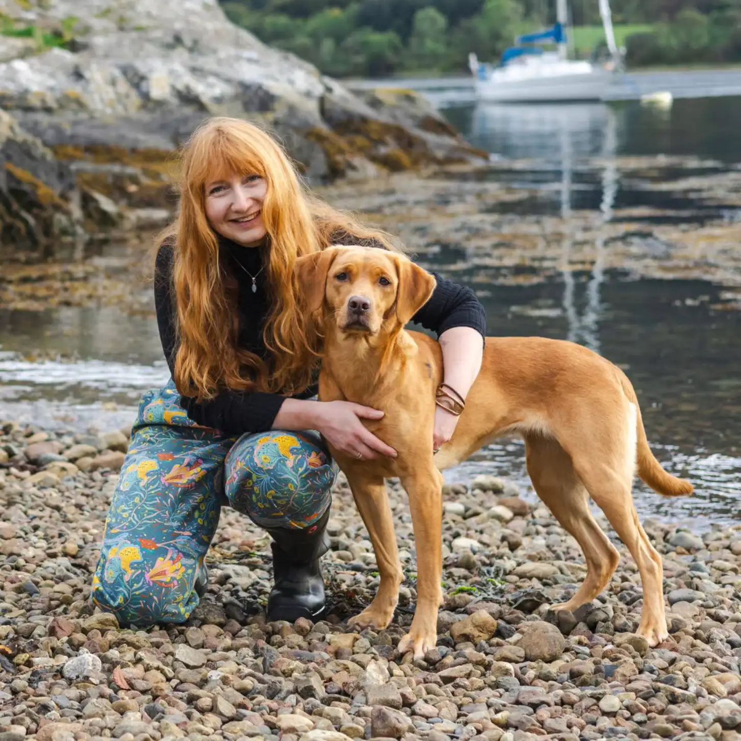 A woman with long reddish-blonde hair crouches on a rocky beach next to a brown Labrador retriever. A sailboat is moored in the water behind them.