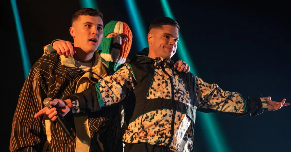 Three performers on stage wearing patterned jackets stand together under blue stage lighting. The center performer wears a ski mask with green, white, and orange stripes. One performer holds a microphone.