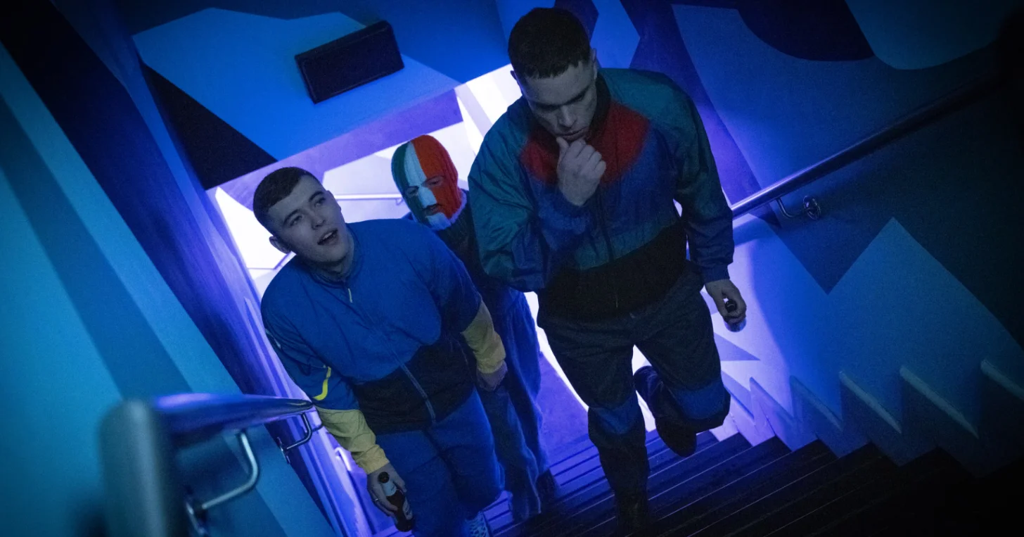 Three people in blue athletic wear stand in a brightly lit corridor with blue and purple neon lighting. The person on the left wears a blue shirt and tan pants, the middle person has a blue, white, and red ski mask, and the person on the right wears a blue and burgundy jacket.