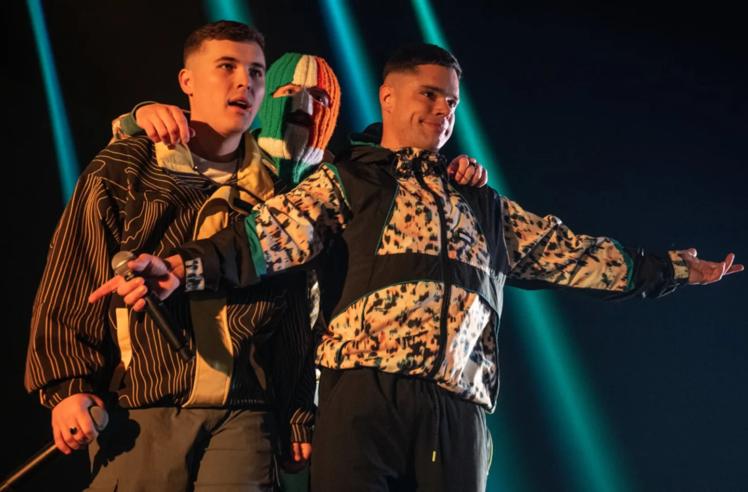 Three performers on stage wearing patterned jackets stand together under blue stage lighting. The center performer wears a ski mask with green, white, and orange stripes. One performer holds a microphone.