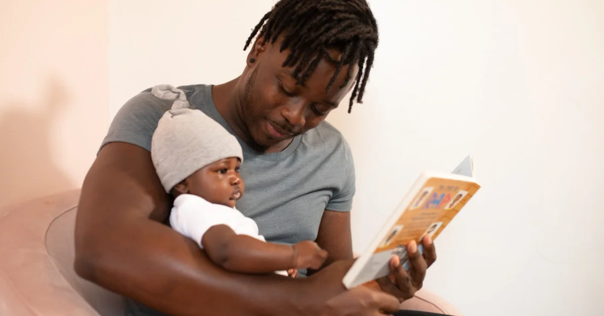 A man holding a baby wearing a white hat looks down at a colorful book they are reading together.