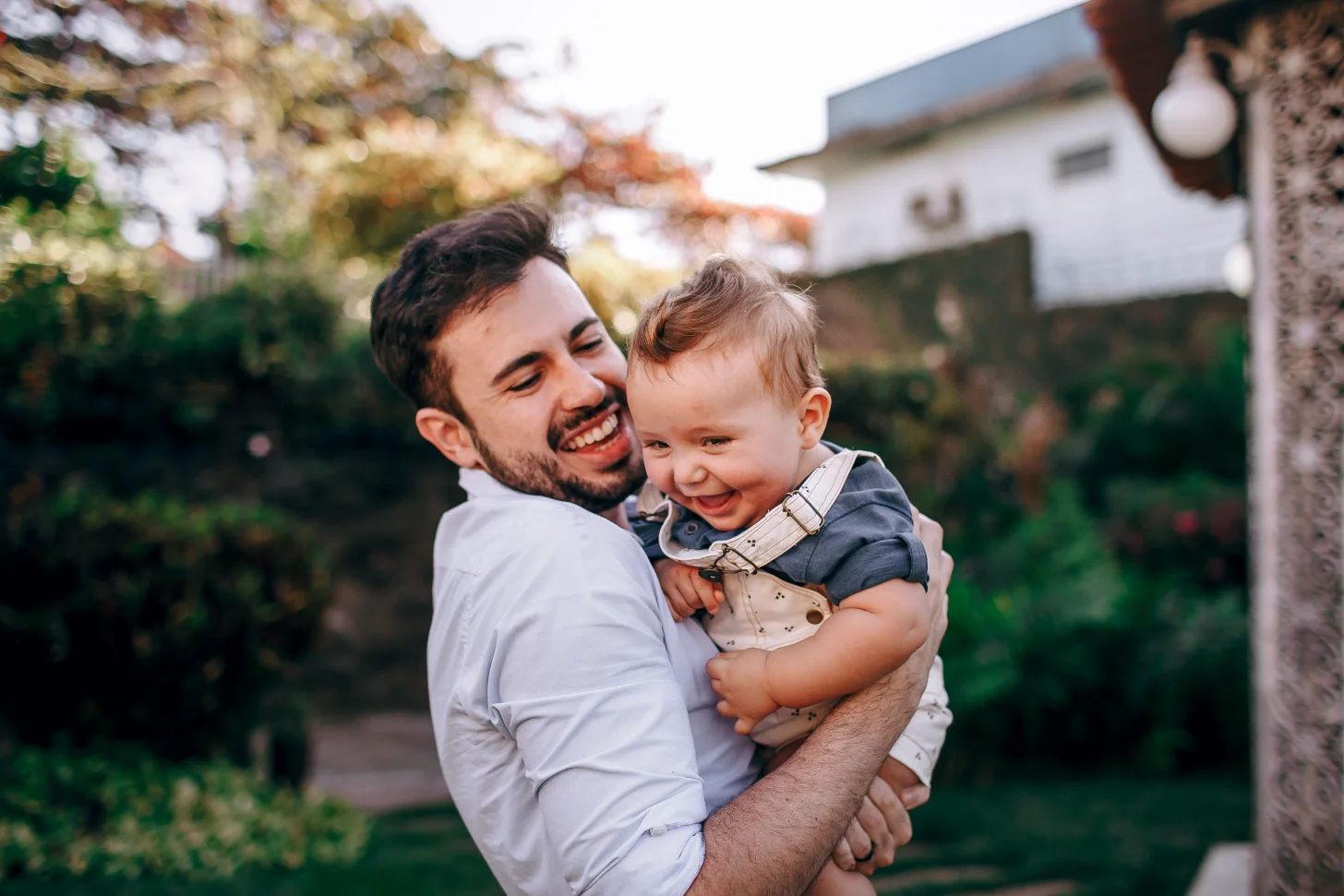 A man in a light blue shirt holds and smiles at a toddler wearing a gray and beige outfit in a residential garden with hedges and a white house visible in the background.