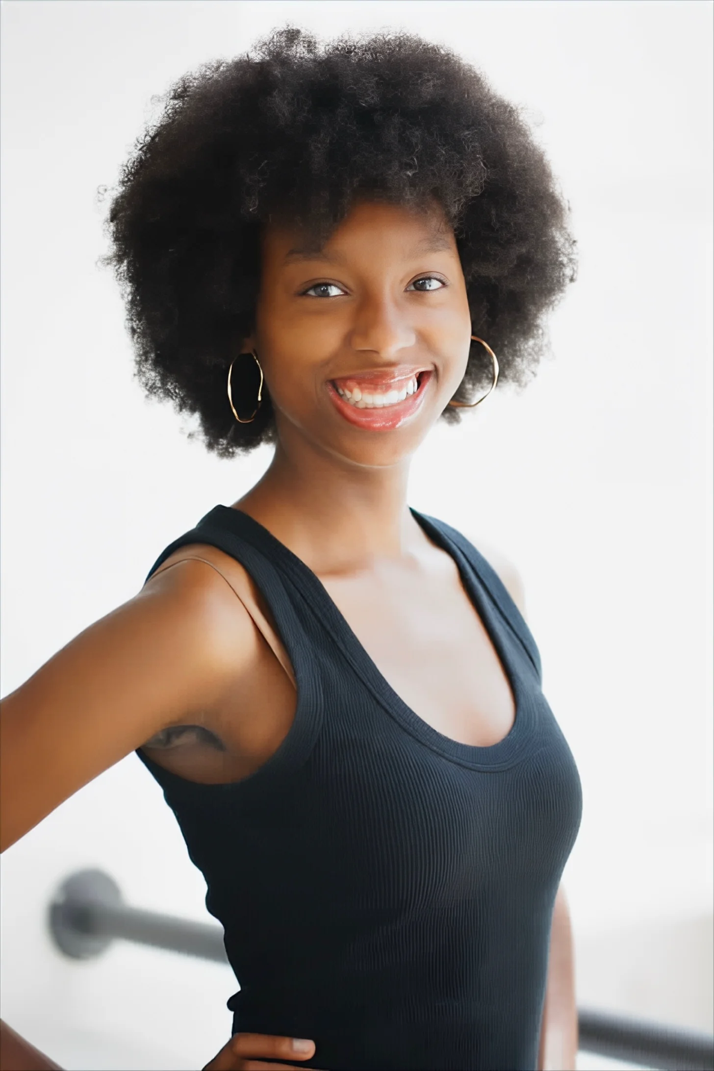 Woman with natural textured hair wearing gold hoop earrings and a dark sleeveless ribbed tank top, smiling at the camera against a white background.
