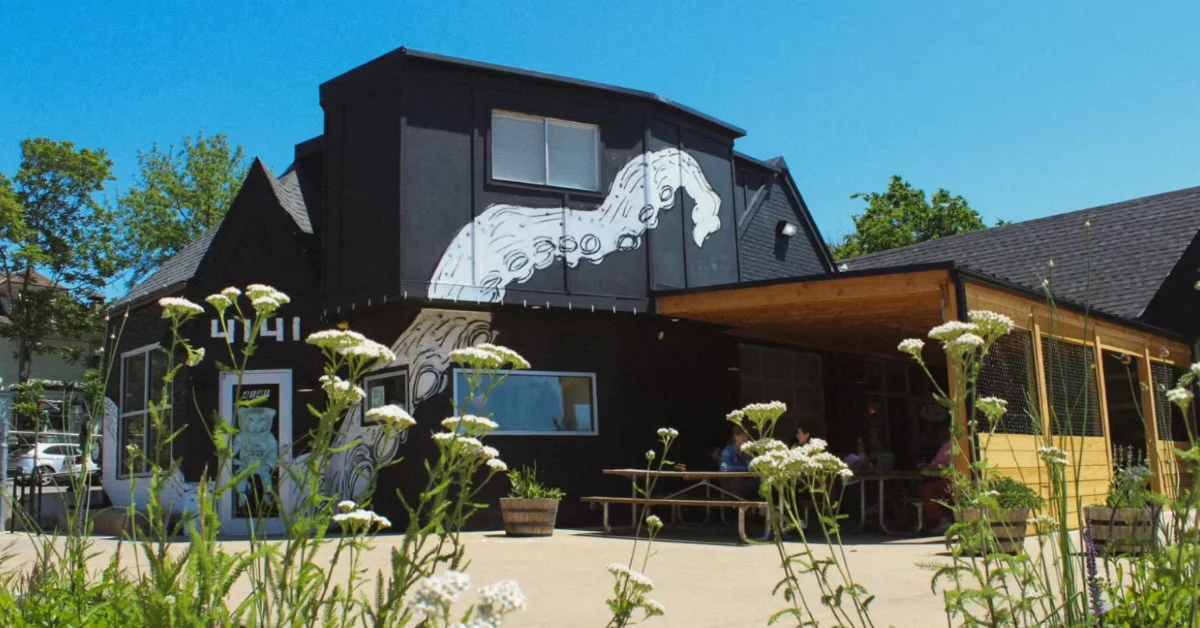 A black building with white octopus mural features wooden structures on either side, white flowering plants in the foreground, and trees against a clear blue sky.
