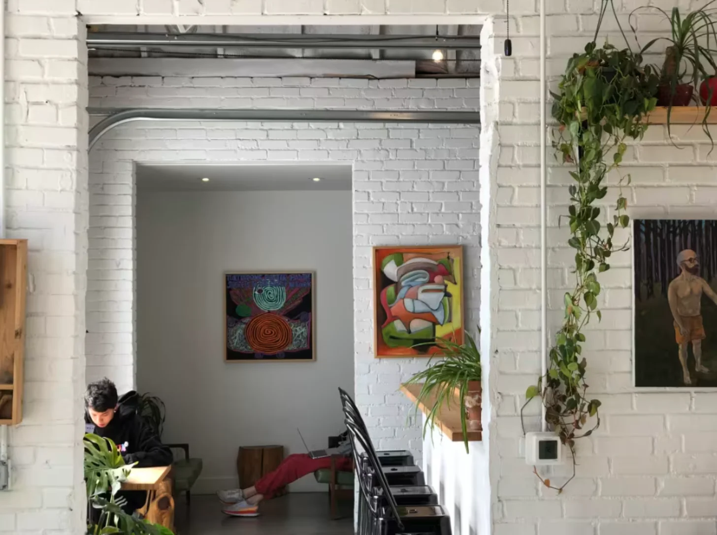 A white brick interior space with a person sitting at a desk on the left side. Colorful artwork hangs on the walls, including abstract paintings with circular and geometric designs. Climbing ivy plants cascade down the right side of the brick wall. A framed portrait of a shirtless man is visible on the far right wall.