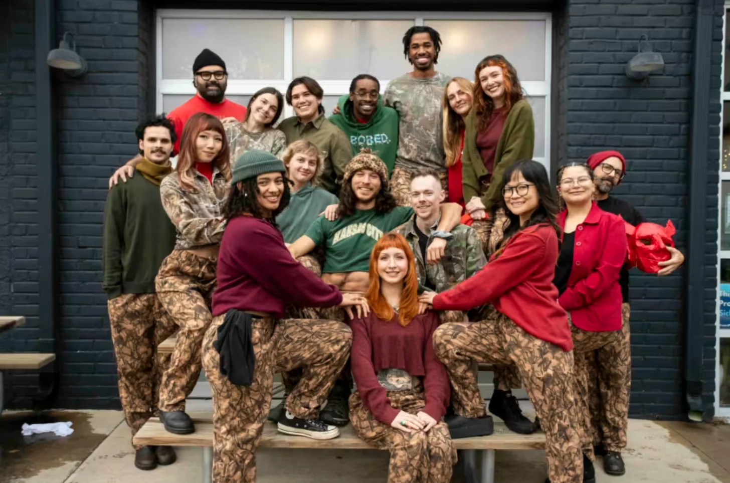 A group of approximately 20 people poses together outside a brick building with large windows, arranged in multiple rows. Many wear camouflage-patterned pants paired with colored tops in red, green, and brown tones.