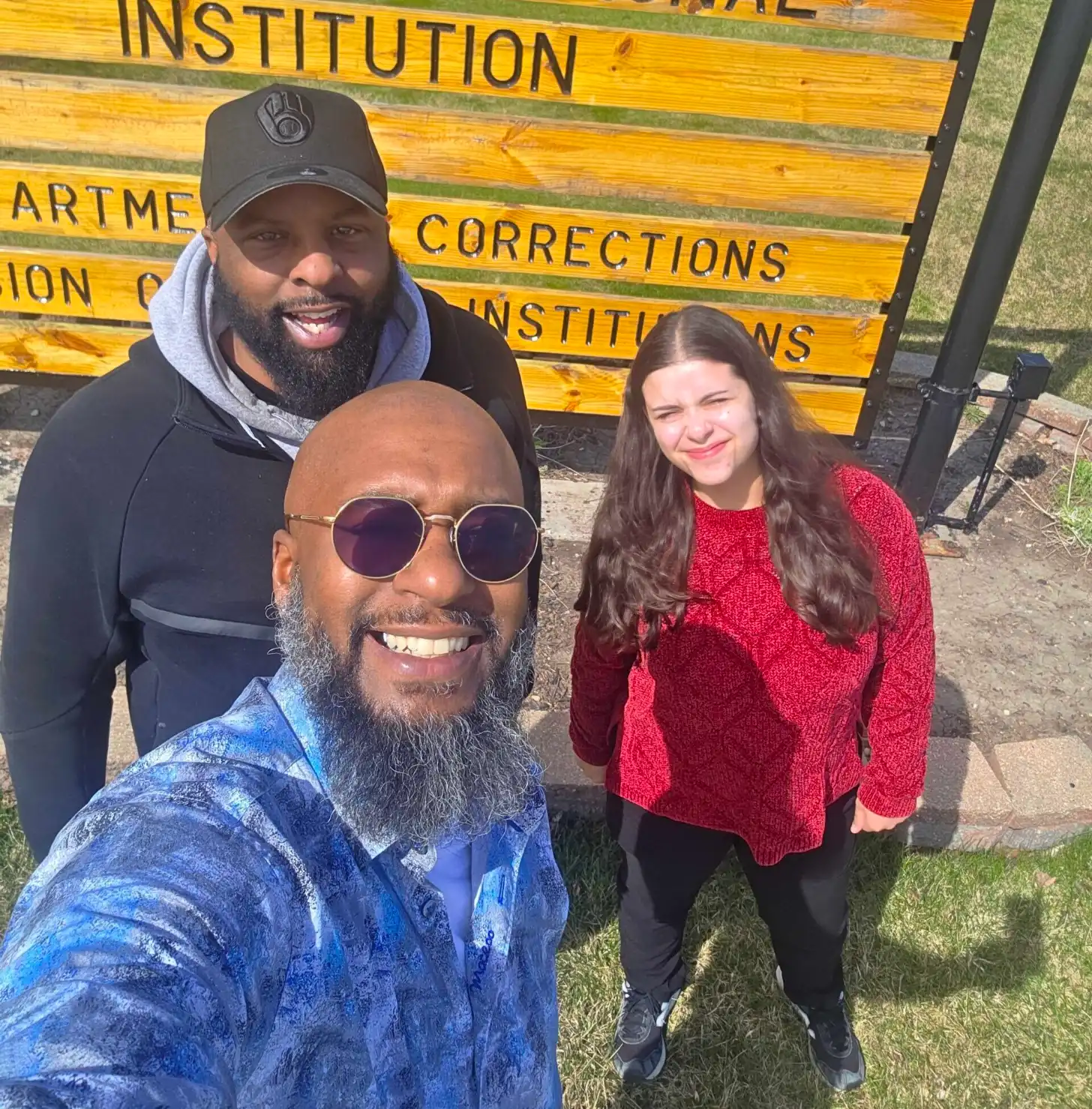 Three people stand outdoors in front of a yellow wooden sign that reads "CORRECTIONS INSTITUTION". The person in front wears a blue jacket and purple-tinted round sunglasses. The person behind wears a black cap and navy hoodie. The person on the right wears a red knit sweater. All three are smiling at the camera.