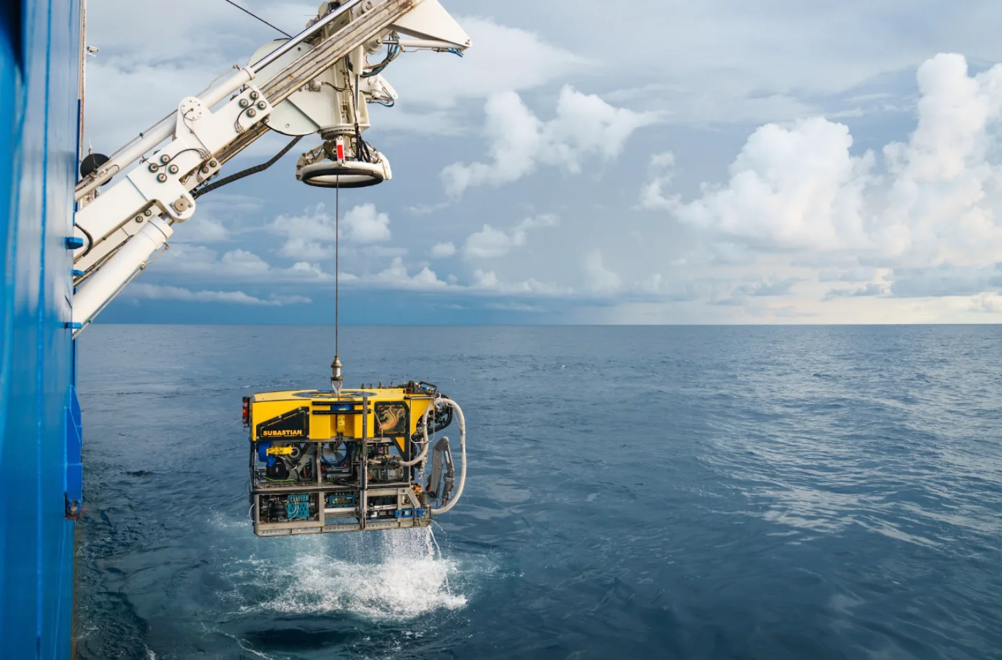 A yellow and blue submersible labeled "SUBASTIAN" is lowered by crane into the ocean from a ship. The device splashes into calm seawater under a partly cloudy sky.