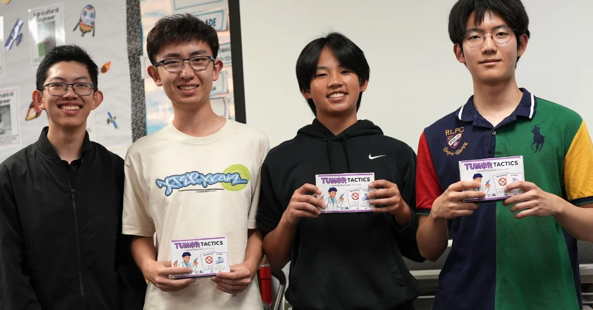Four teenagers hold up copies of "Tumor Tactics" cards while standing in front of a classroom bulletin board decorated with educational posters.