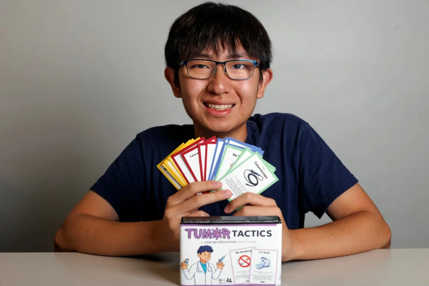 A young person wearing glasses and a dark blue shirt holds fanned-out game cards above a board game box labeled "Tumor Tactics" with cancer education imagery visible on the box.
