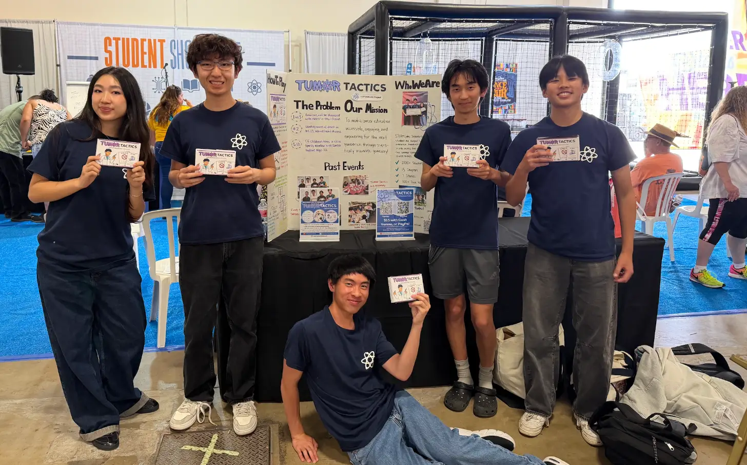 Six students wearing dark blue "Tumor Tactics" t-shirts stand around a display board titled "Tumor Tactics" at a science fair, holding informational cards and smiling at the camera.