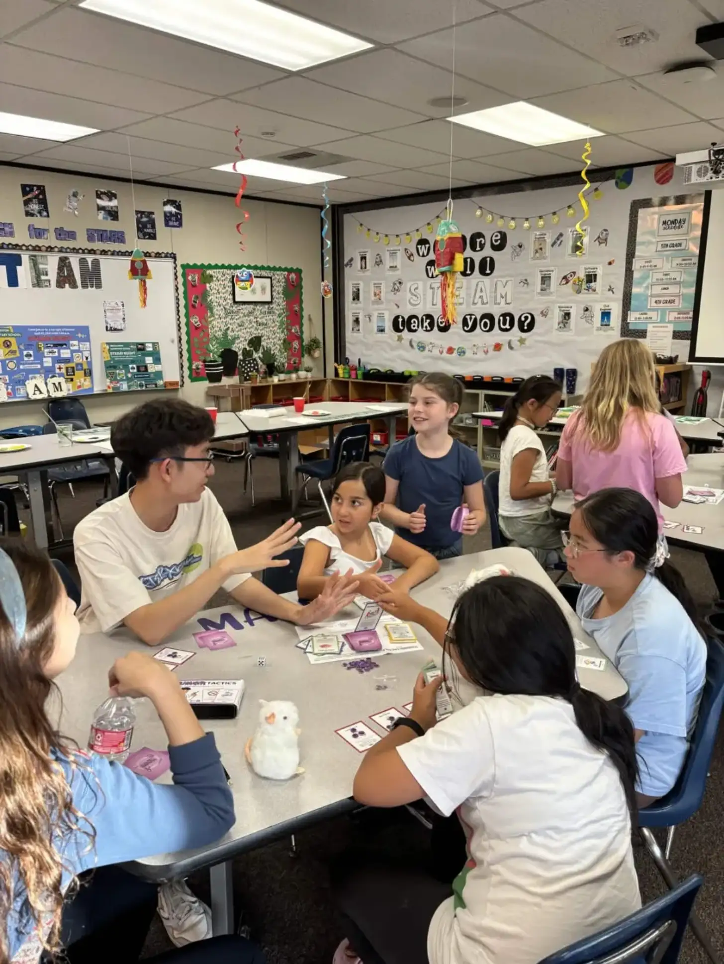Students gather around a table in a classroom playing a card game. A boy in a tan shirt explains rules to the group while others hold cards and listen.