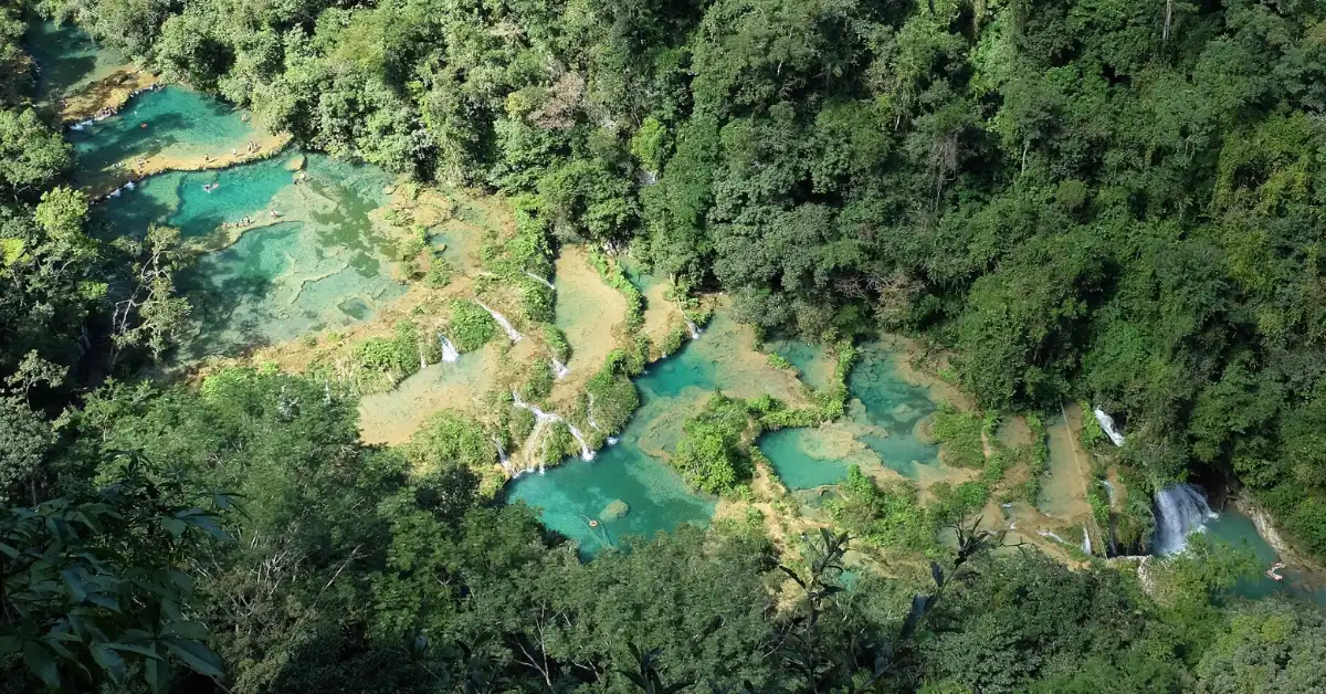 Aerial view of a forested landscape with turquoise and emerald pools of water connected by white waterfalls and winding streams, surrounded by dense green vegetation.