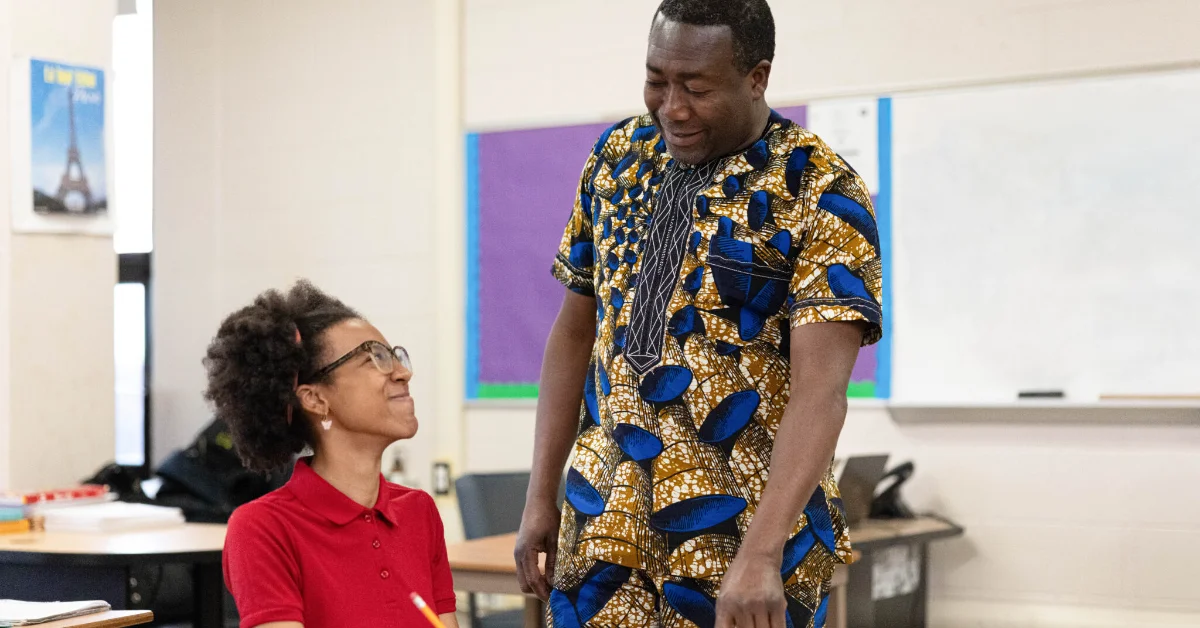 A man wearing a blue and gold patterned shirt stands beside a seated girl in a red polo shirt and glasses in a classroom setting.