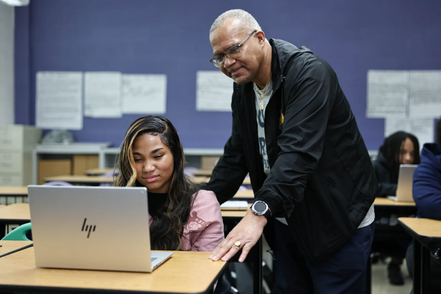 An adult wearing glasses and a black jacket leans over to look at a laptop screen with a student wearing a pink shirt in a classroom setting.