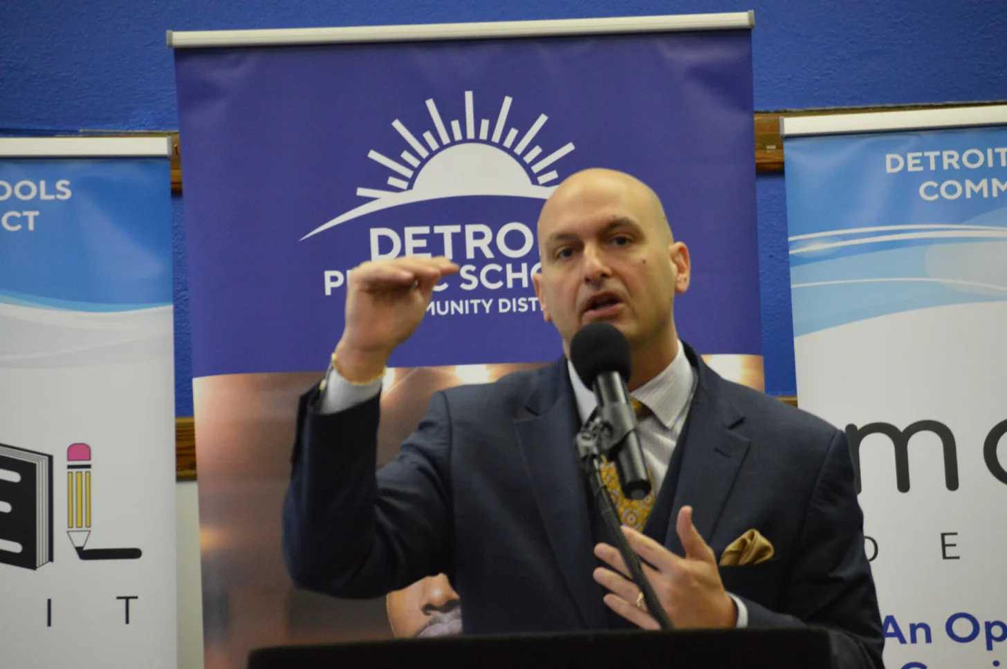 A man in a dark blazer and striped shirt speaks at a microphone with his hand raised, standing before a podium. Behind him is a blue banner with a sun logo and text reading "DETROIT PUBLIC SCHOOLS COMMUNITY DIST."