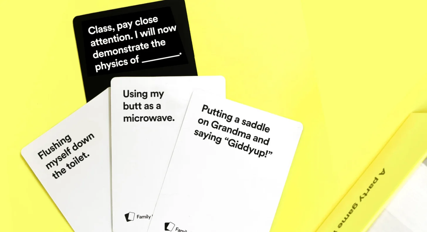 Cards spread on a yellow background with text prompts from a game. Visible cards read: "Class, pay close attention. I will now demonstrate the physics of ___," "Using my butt as a microwave," "Putting a saddle on Grandma and saying 'Giddyup!'," and "Flushing myself down the toilet."