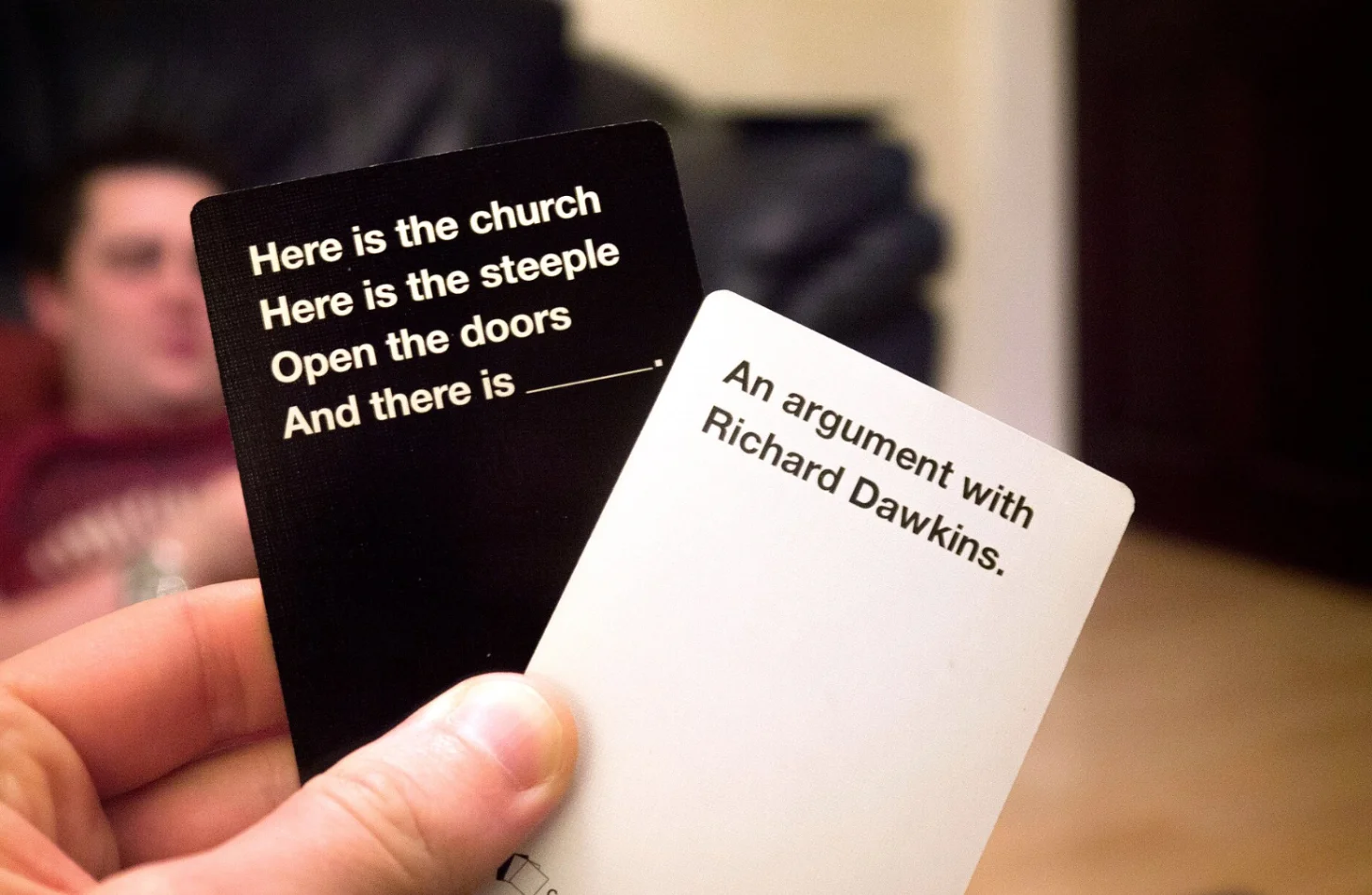 A hand holds two cards from a game. The black card reads "Here is the church / Here is the steeple / Open the doors / And there is ___." The white card reads "An argument with Richard Dawkins."