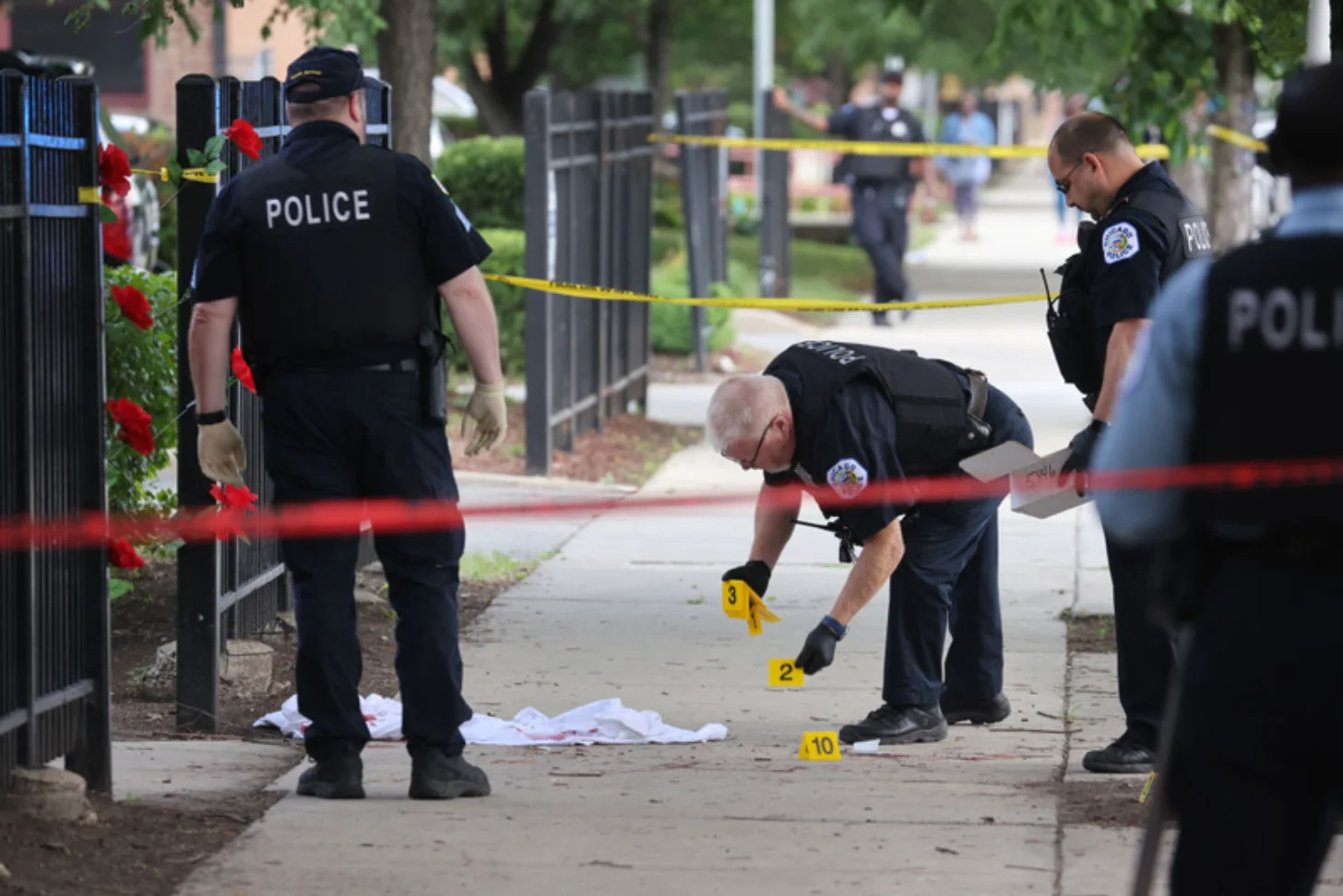 Police officers place evidence markers on a sidewalk at a crime scene cordoned with yellow tape, with red flowers visible on a fence nearby.