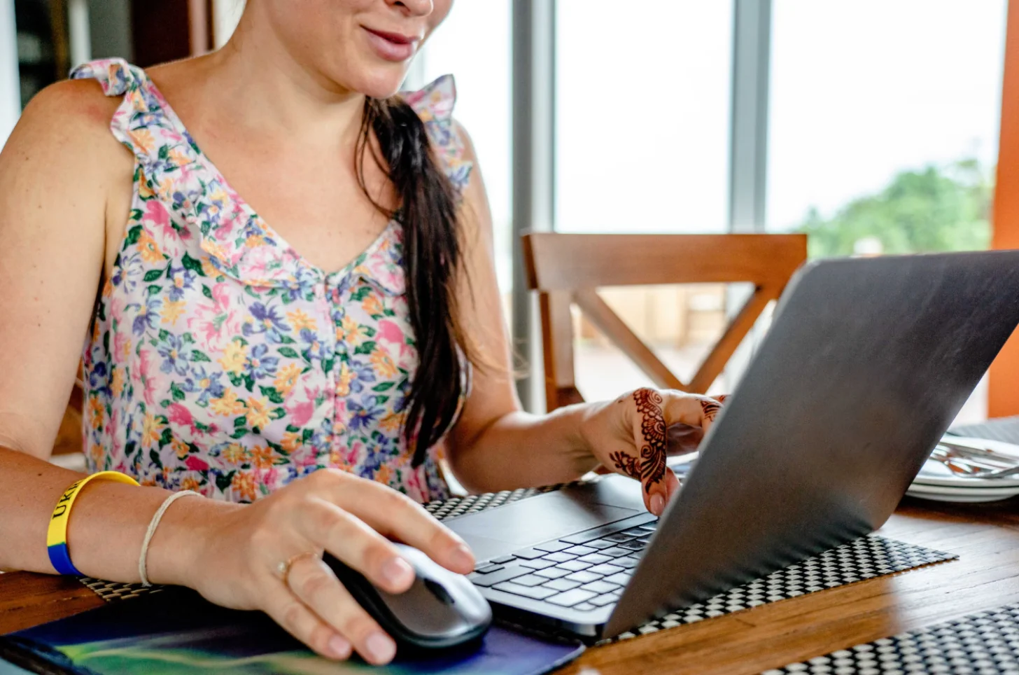 Woman wearing a colorful floral tank top uses a laptop and mouse at a wooden table, with a wooden chair and windows visible in the background.