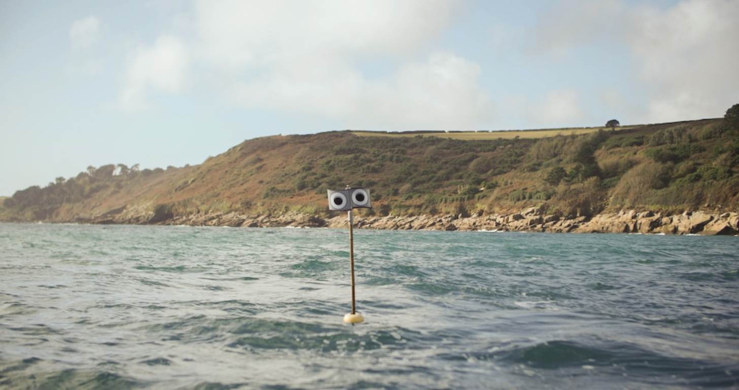 A pole with two circular speakers mounted on top stands in shallow ocean water, with rocky cliffs and green hillside visible in the background.