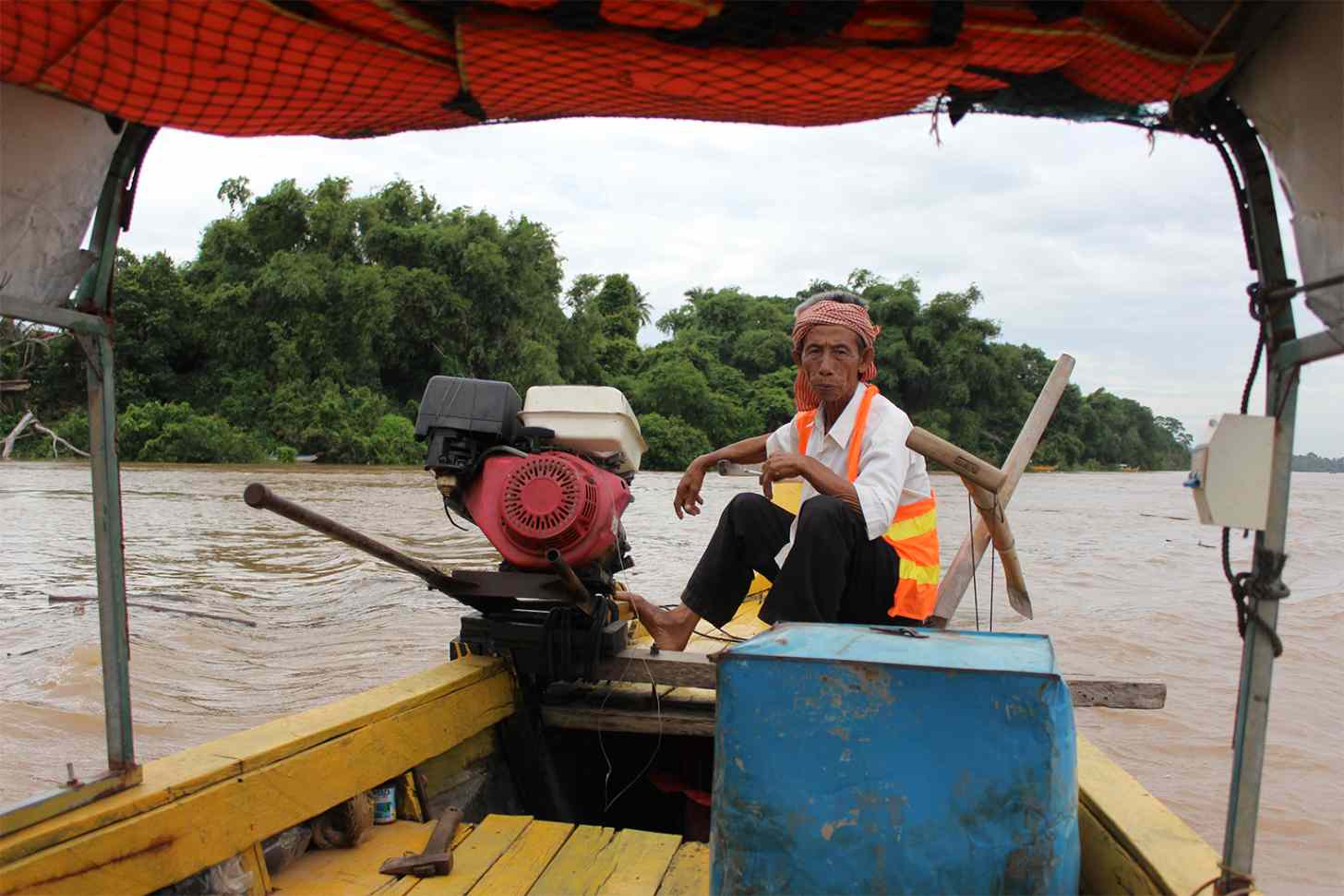 A man wearing a red checkered head covering and orange safety vest sits in a yellow boat on a brown river, operating a red outboard motor.