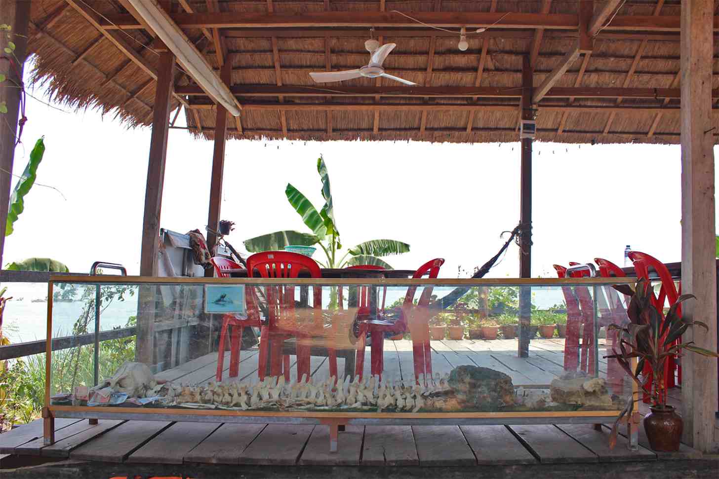 Red wooden chairs and a table beneath a thatched roof structure, with a glass display case containing small figurines in the foreground and banana plants visible in the background.