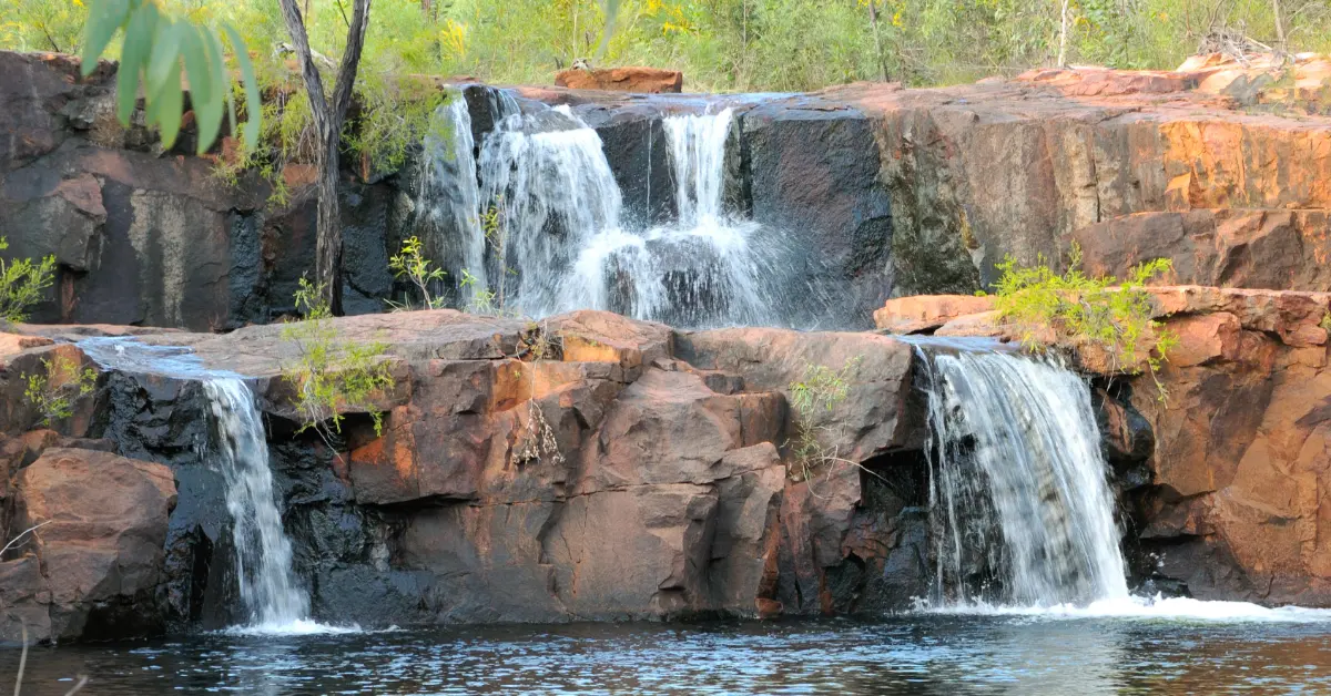 Multiple tiered waterfalls cascade over reddish-brown rocky outcrops into a dark pool below, surrounded by green vegetation and trees.