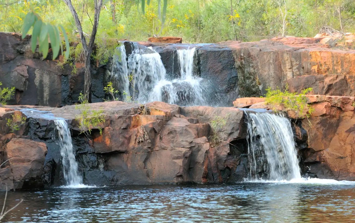 Multiple waterfalls cascade down layered reddish-brown rock formations into a pool of water. Green vegetation and trees surround the rocky terrain in the background.
