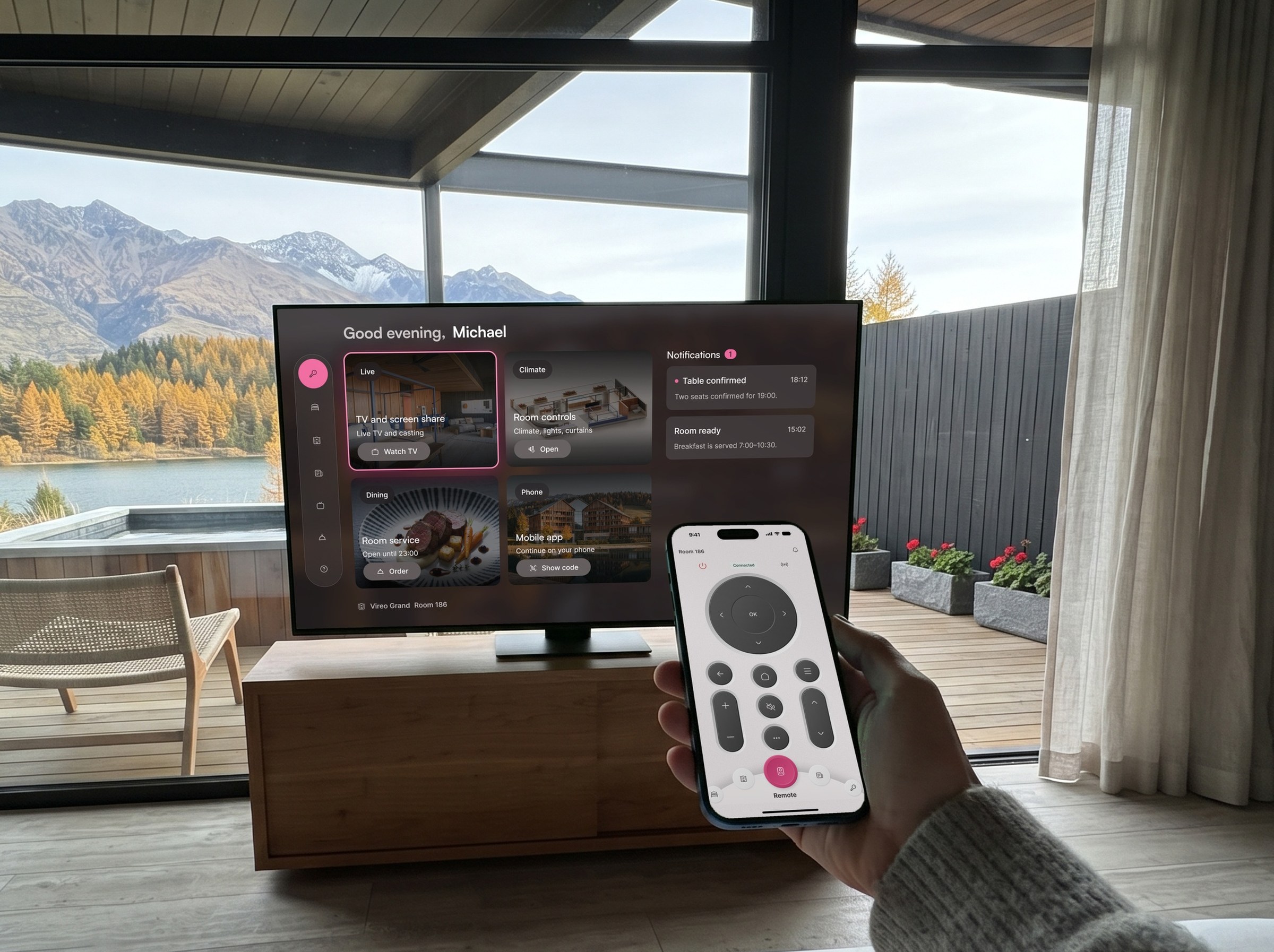 A guest on the edge of the bed holds a phone with the remote open, pointed at the room TV showing the Roomside home screen.