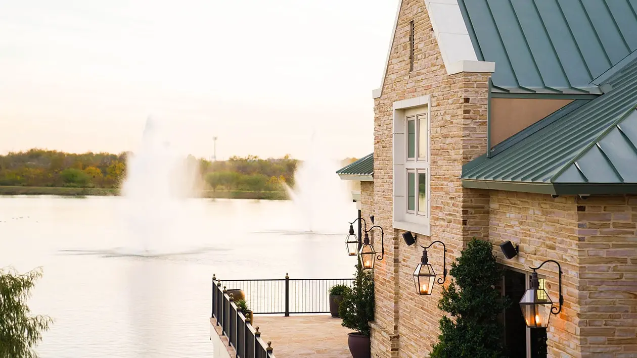 Charming lakeside view of a stone building with green metal roof at sunset. Lit lanterns adorn the balcony, overlooking fountains in a calm lake. The Red Berry Estate in San Antonio Texas