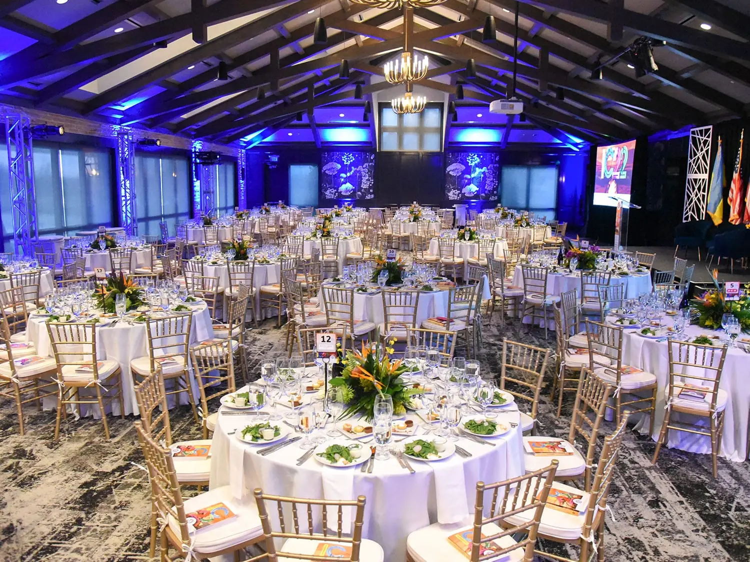 Elegant banquet hall with round tables set for an event. Tables have white tablecloths, place settings, and floral centerpieces. Blue ambient lighting creates a formal atmosphere.