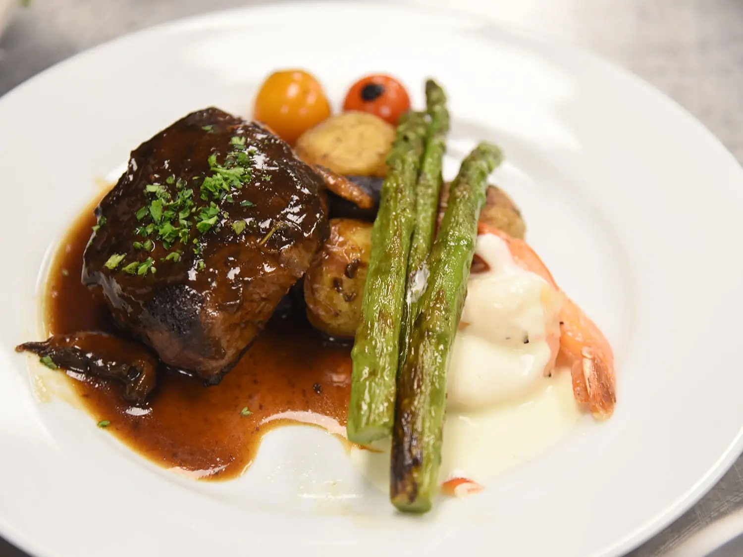 A gourmet meal features a glazed steak topped with herbs, asparagus, roasted potatoes, cherry tomatoes, and creamy sauce on a white plate, exuding elegance.