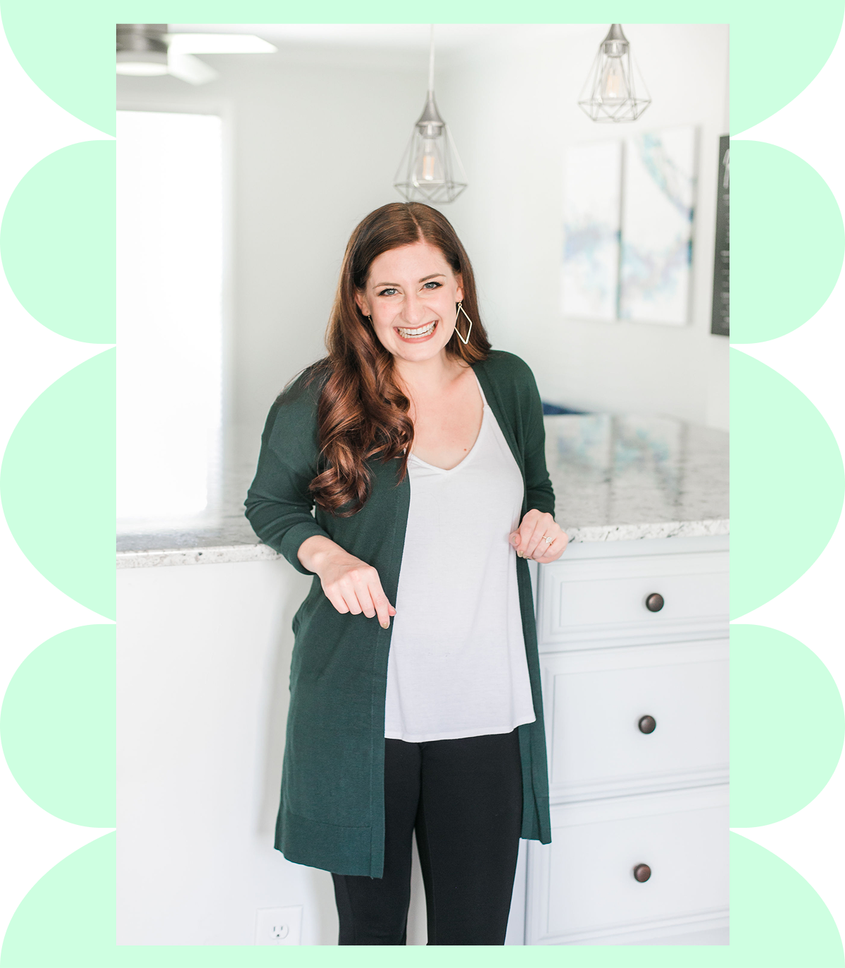 Smiling woman with long brown hair wearing a dark green cardigan, white top, and black pants standing in a bright kitchen.