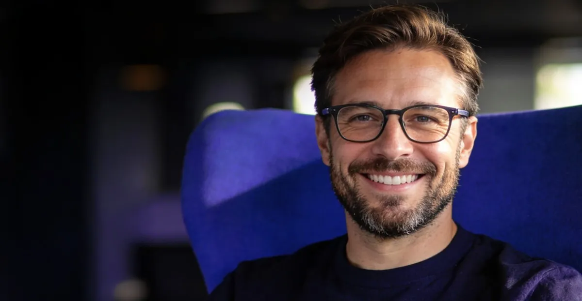 Man with glasses and beard smiling while sitting in a blue chair.