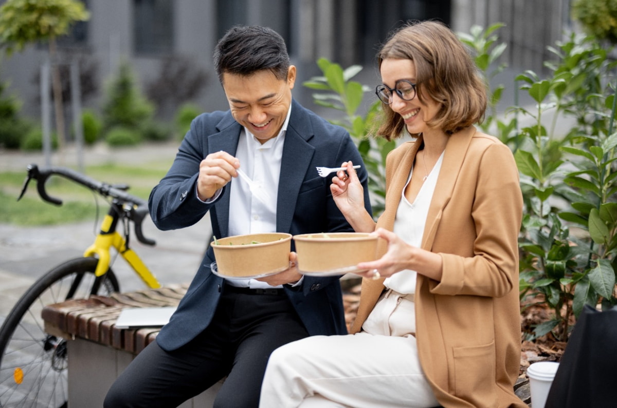 A couple enjoys a meal outdoors, sitting on a bench, with a bicycle nearby, surrounded by greenery.