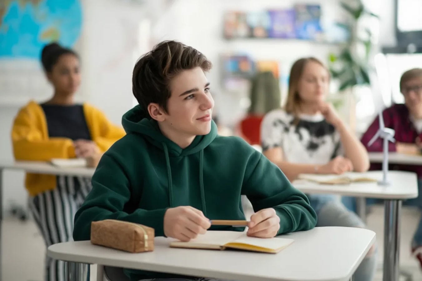 Student in a green hoodie attentively listens in a bright classroom, with classmates and world maps in the background, conveying engagement and focus.