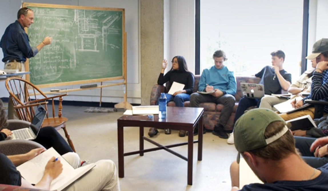 students at Middlebury College gathered around in chairs and sofas as a professor at a chalkboard explains a complex topic