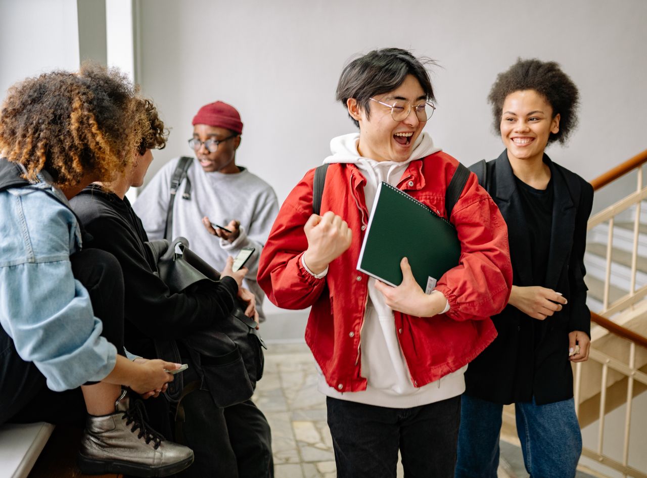 student holding a notebook celebrates with his friends around him in a campus stairwell leaving class
