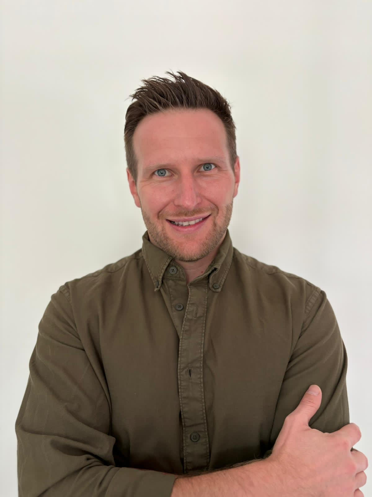 Smiling man with short brown hair and blue eyes wearing an olive green button-up shirt with arms crossed.