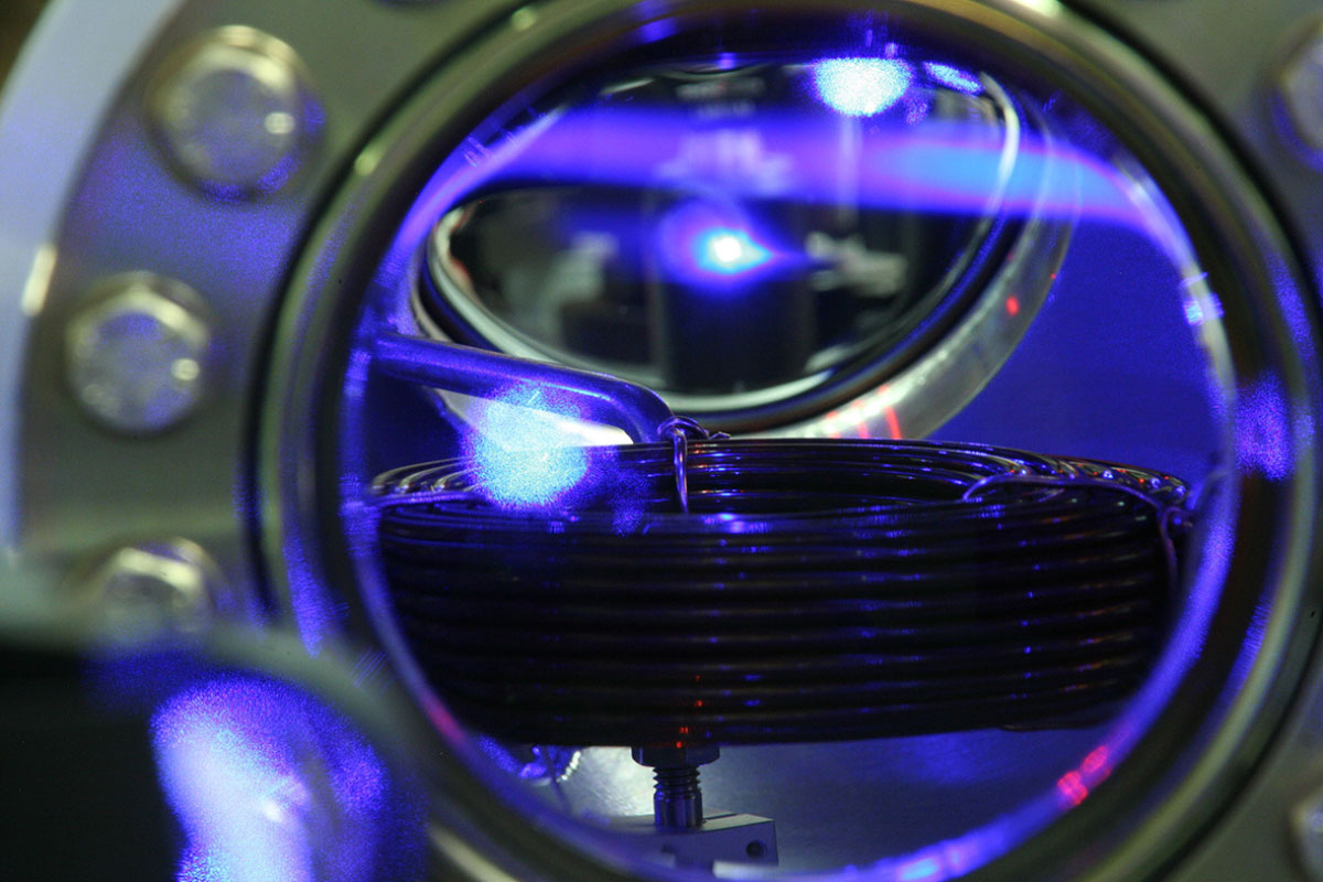 View of the ultra-high vacuum chamber of PTB's optical strontium clock where strontium atoms are cooled and stored. The brilliant blue/white fluorescent light in the upper third of the window results from a cloud of cold strontium atoms (the drop-shaped formation below the blue fluorescent atom beam in the upper part of the vacuum window).