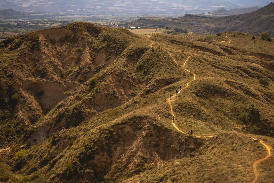 A winding dirt path on dry, grassy hills with five mountain bikers riding in a natural landscape.