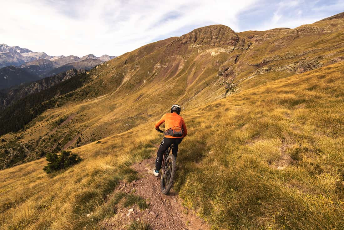 Cyclist wearing an orange jacket riding a mountain bike on a narrow trail in grassy hills.