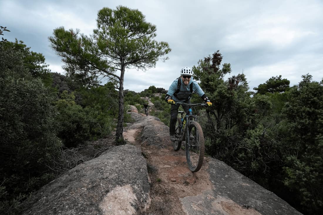 Man wearing helmet mountain biking on rocky trail surrounded by trees under cloudy sky.