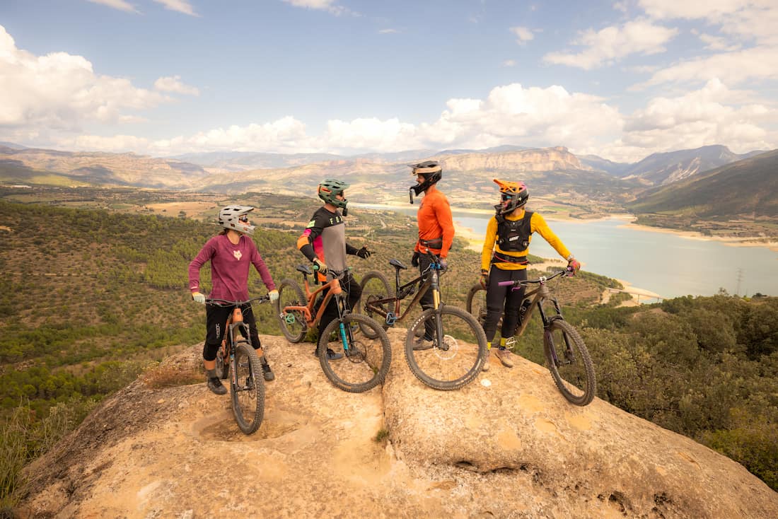 Four mountain bikers wearing helmets standing with bikes on a rocky overlook over valley and lake.