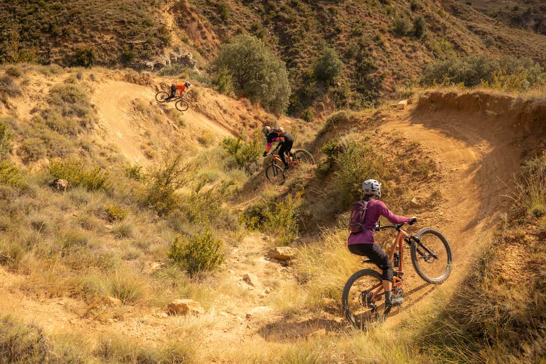 Three mountain bikers riding on a dirt trail winding through dry, hilly terrain with scattered bushes.