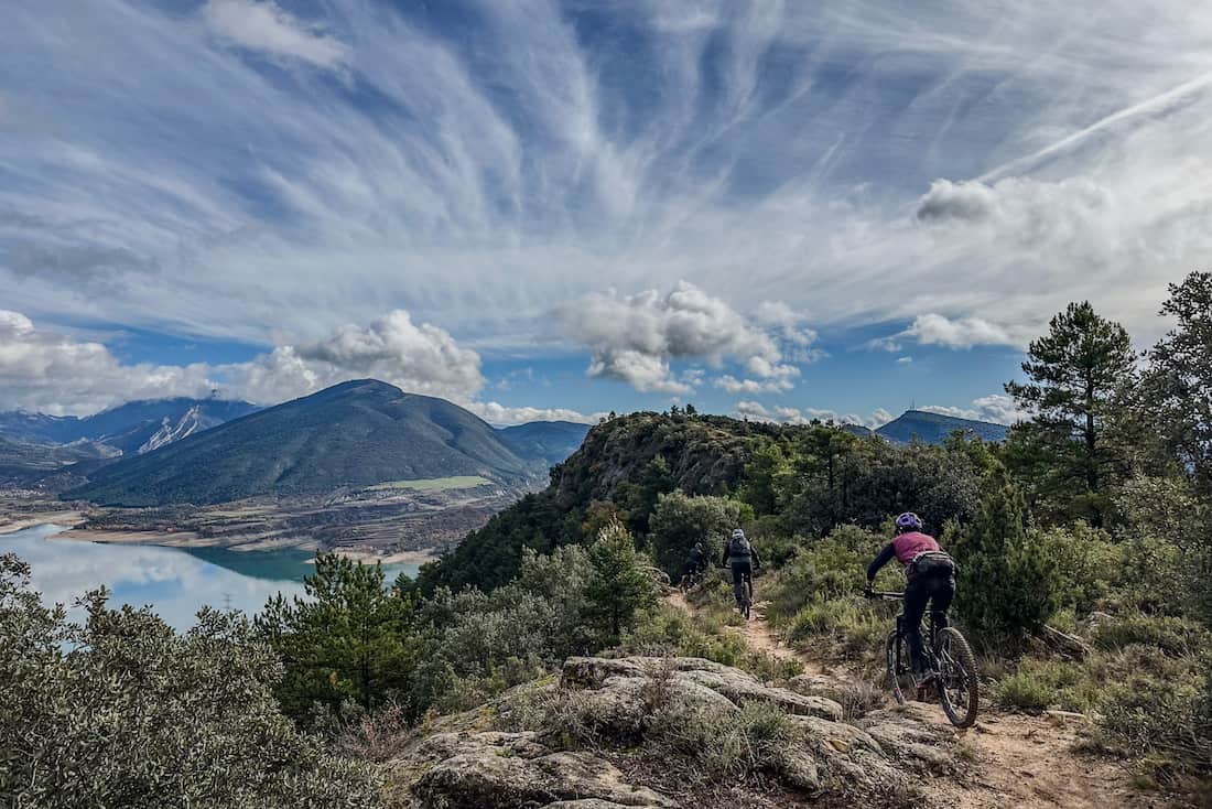 Three cyclists ride on a mountain trail surrounded by trees with a lake and hills in the background.