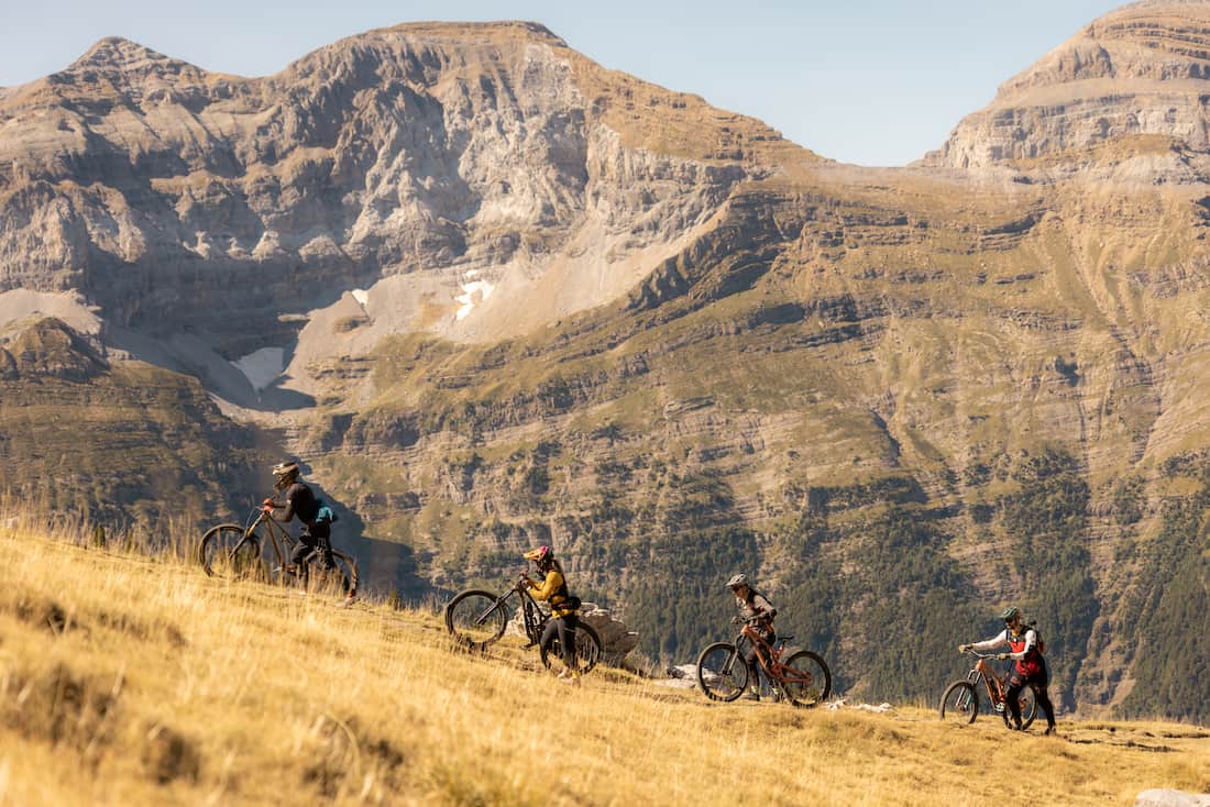 Four people pushing mountain bikes uphill on grassy slope with rocky mountains in the background.