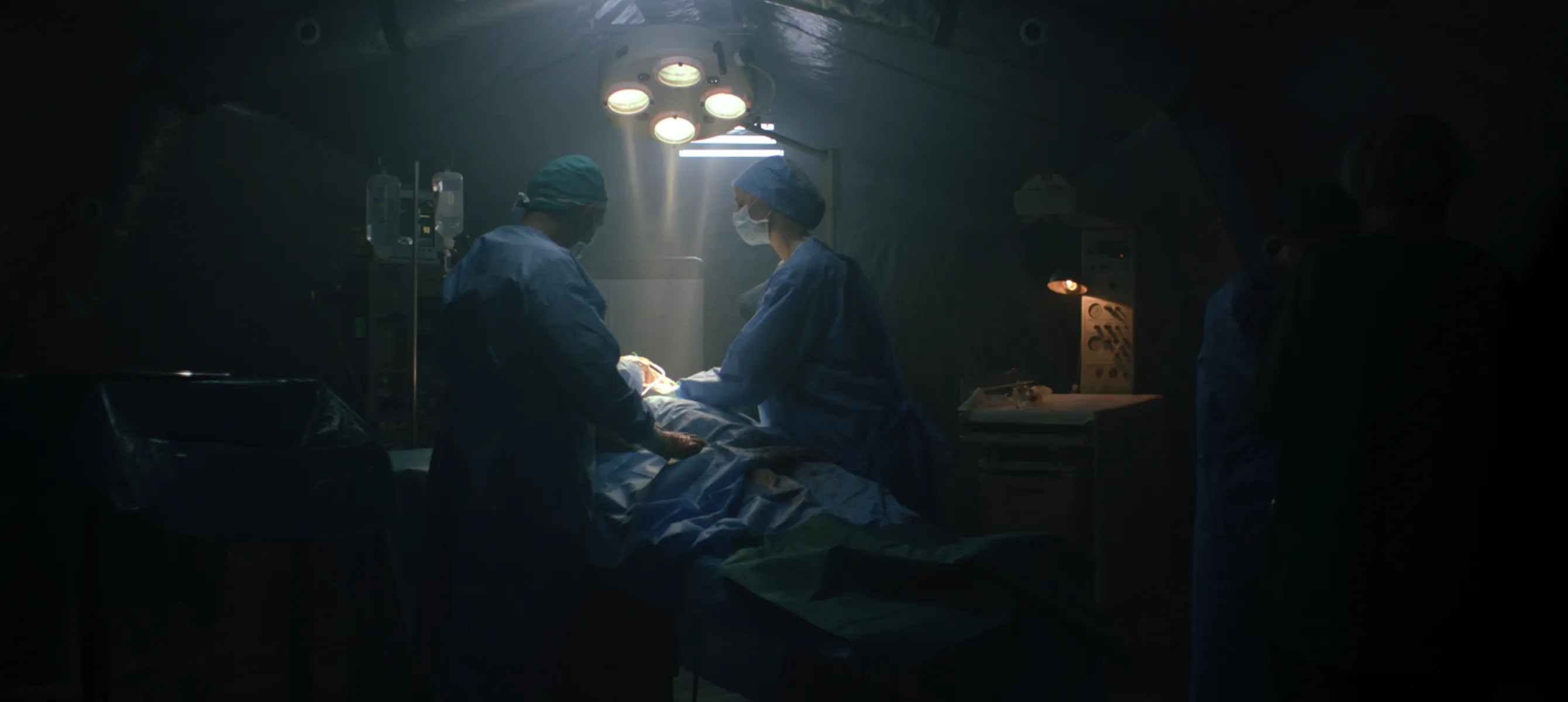 Two surgeons in blue scrubs performing surgery under bright overhead lights in a dimly lit operating room.