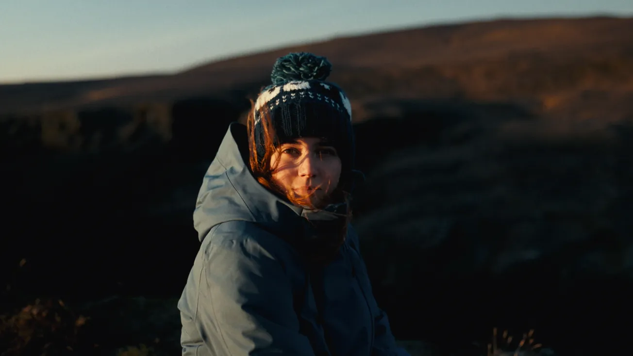 Young woman wearing a knitted hat with a pom-pom and a blue jacket, outdoors near rocky terrain at dusk.